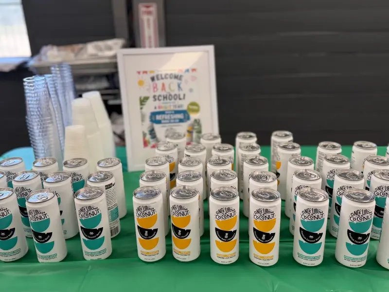 Rows of coconut water cans and stacks of cups on the green-clothed drinks table, a framed welcome-back sign behind them