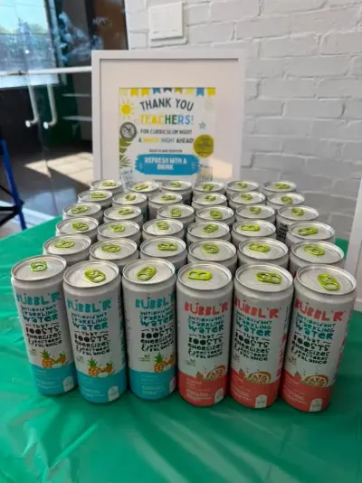 Rows of Bubbl’r sparkling water cans on a green table in front of a framed ‘Thank you teachers, for Curriculum Night’ sign