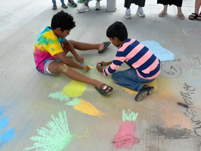 Two children sitting on the concrete filling in a patch of chalk colour, handprints and drawings spreading around them