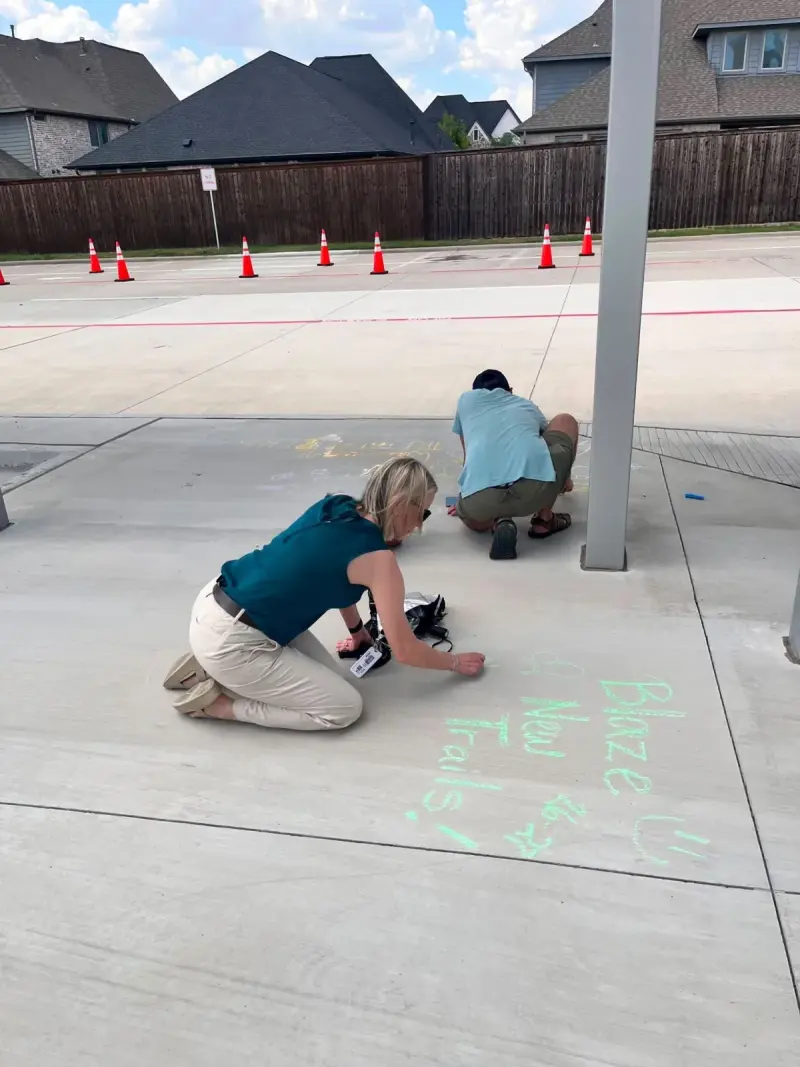 Two volunteers kneeling on the walkway to chalk a message in green, traffic cones lined up across the car park behind