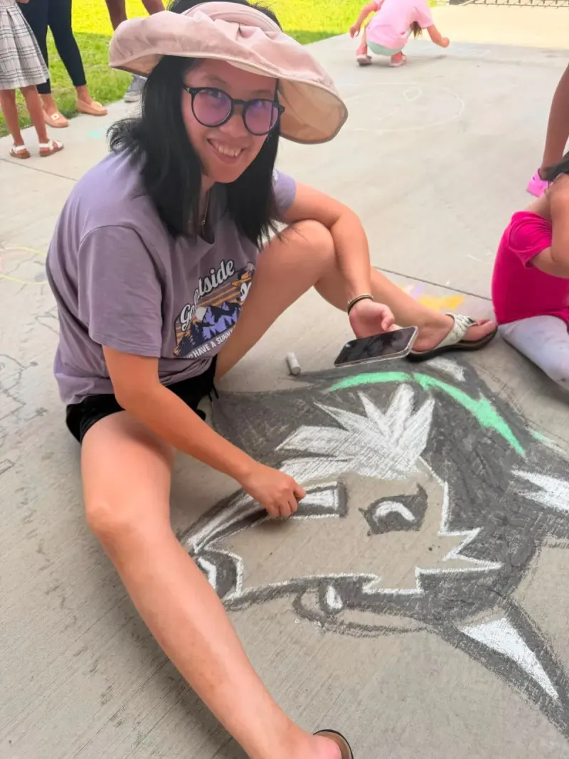 A student in a wide sun hat sitting back from the husky head she has been drawing, phone in hand for reference