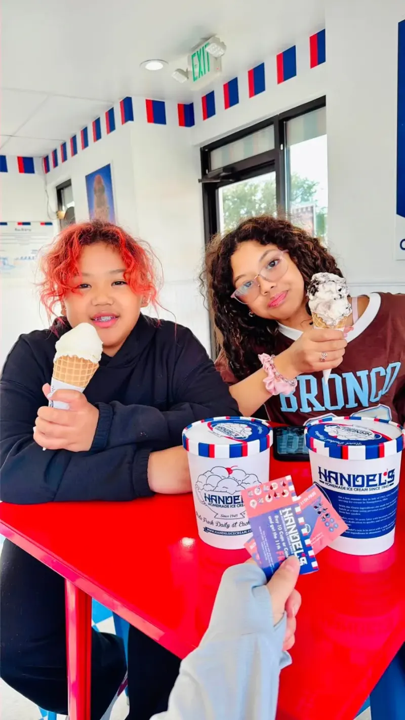 Two children at a red table in the ice cream parlour holding up cones, tubs and spirit night vouchers in front of them