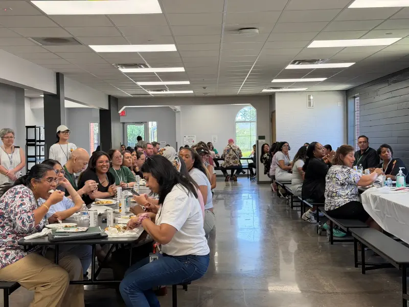 The length of the cafeteria at lunch, staff on both sides of the aisle eating and talking