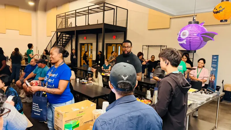 Teachers and staff chatting across the cafeteria with plates of breakfast, a purple paper bat lantern hanging overhead