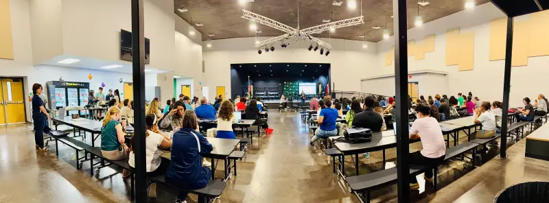 A panoramic view of the cafeteria, teachers and staff filling the long tables with the stage at the far end