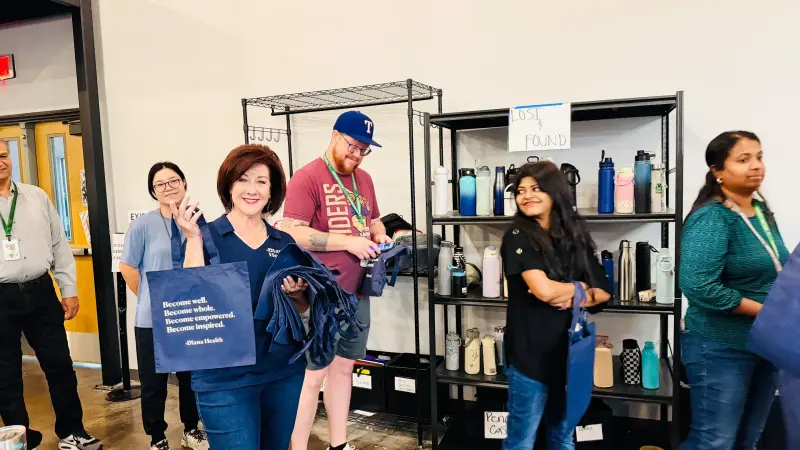 The Diana Health representative holding up navy tote bags printed “Become well. Become whole. Become empowered. Become inspired.” as staff gather by the lost and found