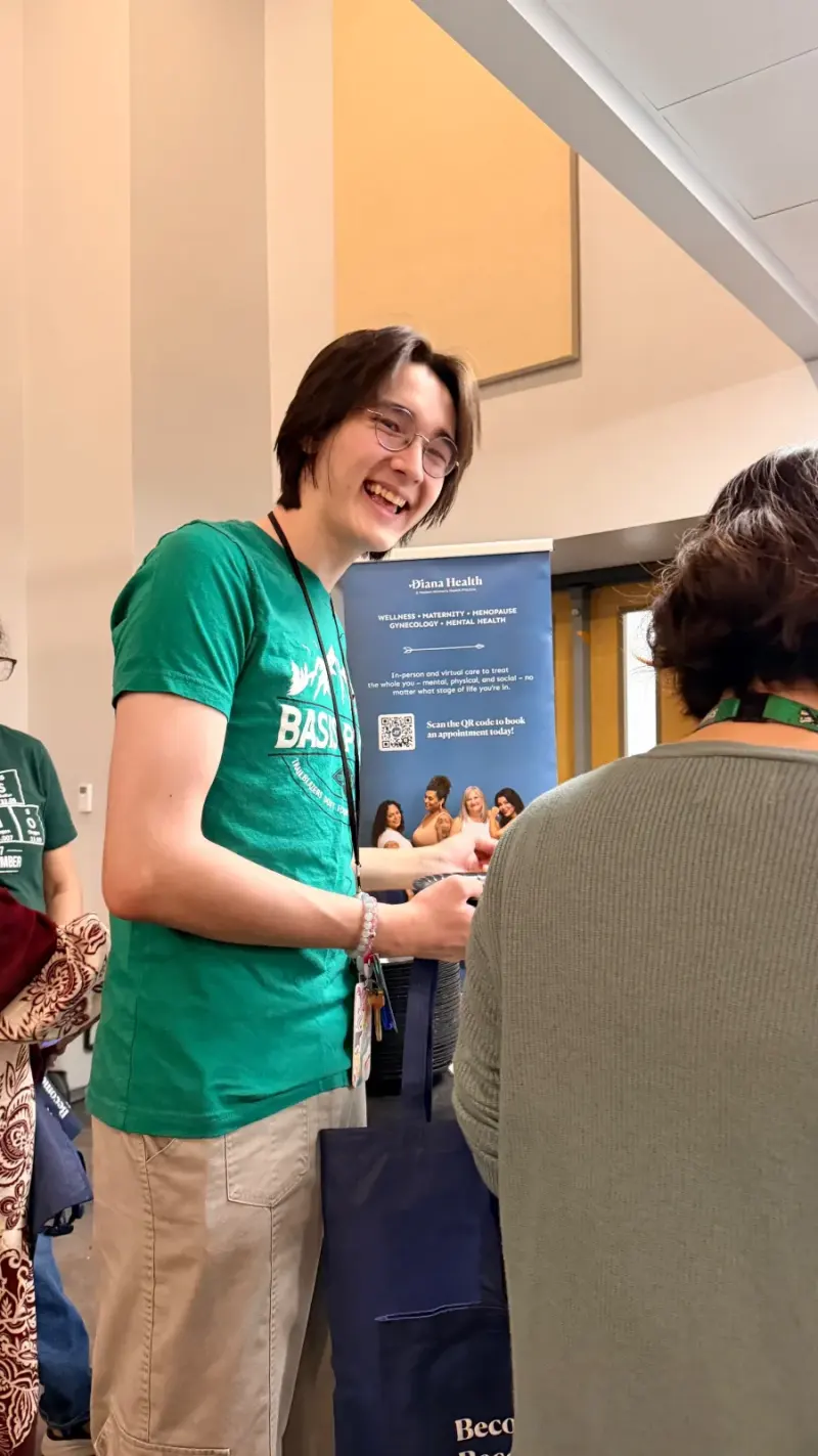 A smiling staff member in a green BASIS shirt collecting a Diana Health tote bag beside the Diana Health banner