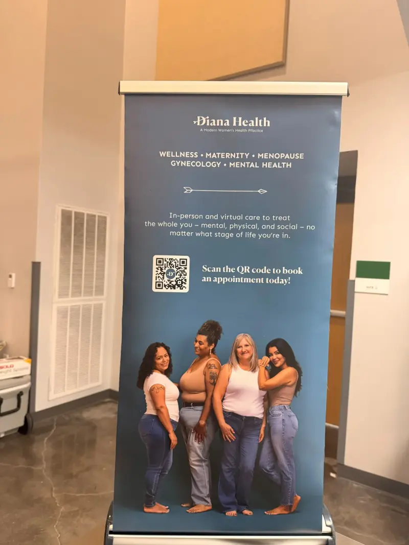 Diana Health’s pull-up banner in the cafeteria — wellness, maternity, menopause, gynecology and mental health care, with a QR code to book an appointment