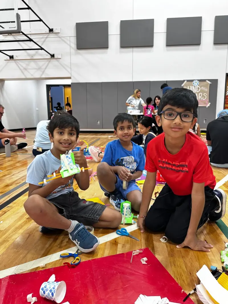 Three boys on the gym floor with their builds, one holding up a paper-cup wind car on straw axles, the “Wind Cars” sign behind them