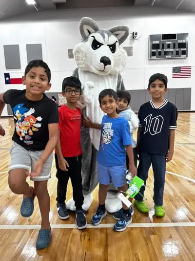 Five boys grinning with Blaze the Husky mascot in the gym, one holding his paper-cup wind car