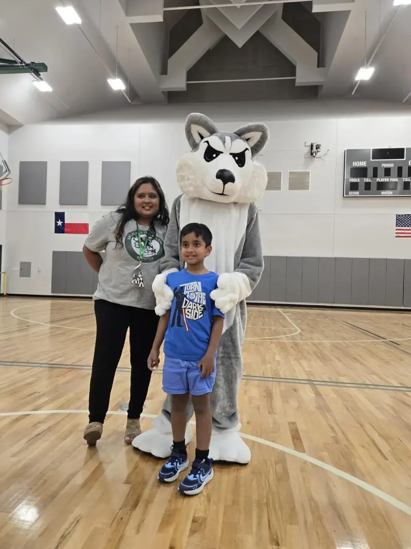 A Booster volunteer in a Trailblazers T-shirt and a boy in a blue T-shirt posing with Blaze the Husky mascot on the gym floor