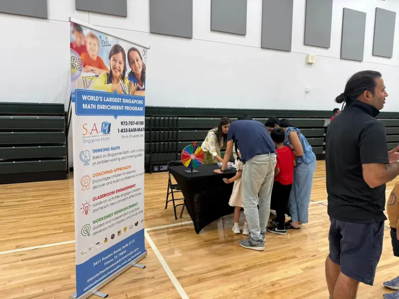 Families leaning over the Singapore Math table beside a spinning prize wheel, the program’s tall banner standing in the foreground