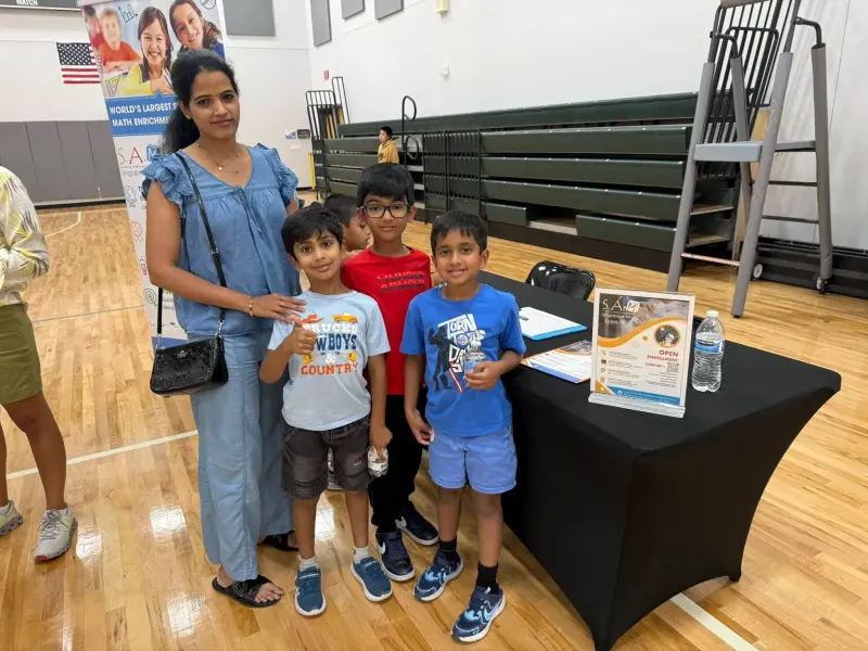 A parent and four boys smiling beside the Singapore Math table, an open-enrollment flyer and sign-up sheet on the black tablecloth