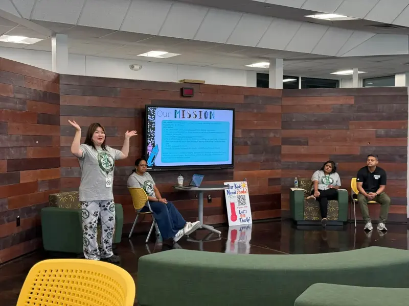 The General Meeting in the lounge: a Booster volunteer opening with arms wide beside an “Our Mission” slide, the rest of the team seated along the wood-panelled wall