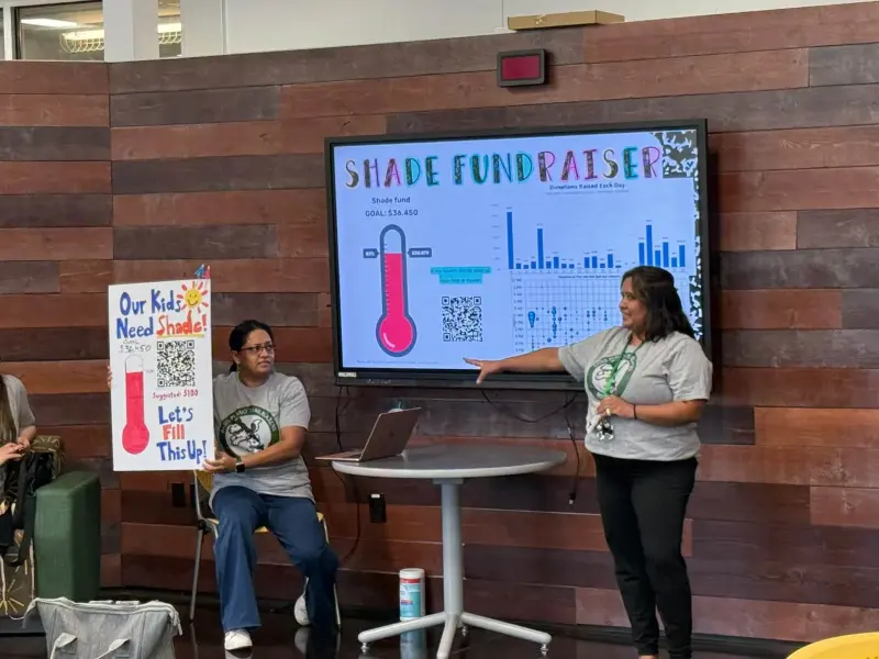 A Booster volunteer pointing to the shade fundraiser’s progress thermometer on screen while another holds up the hand-drawn “Our Kids Need Shade!” poster