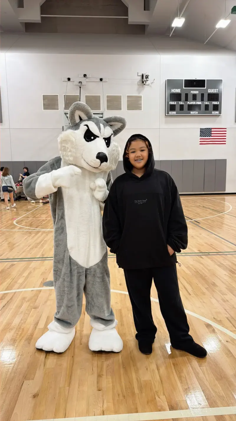 A student in a black hoodie smiling beside Blaze the Husky mascot on the gym floor, the scoreboard and the American flag behind them