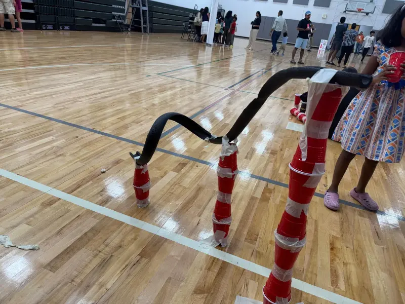 A roller coaster track of black foam tubing taped onto towers of stacked red cups, built on the gym floor