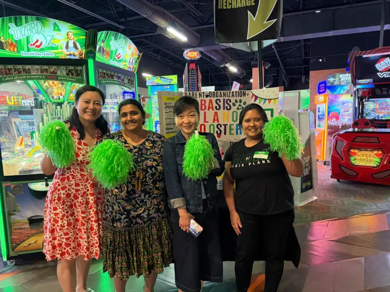 Four Booster volunteers holding green pom-poms in front of the arcade games, the display board behind them