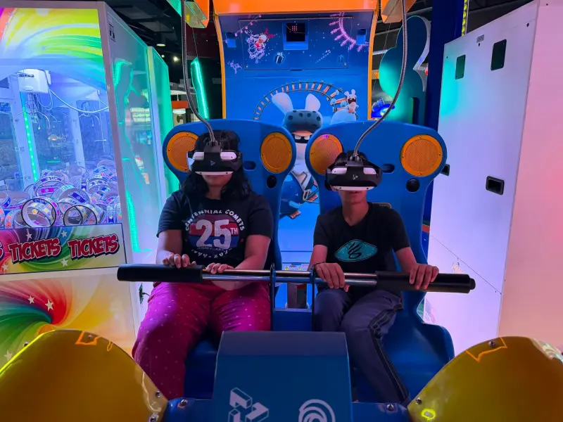 Two children strapped into a virtual reality ride wearing headsets, hands on the safety bar