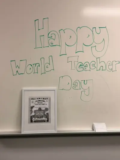 “Happy World Teacher Day” drawn in green bubble letters on a whiteboard, above a framed Booster flyer: “Celebrating World Teacher Day! A special present for all teachers and staff, from the BASIS Plano Booster”
